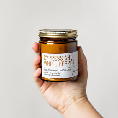 Hand holding a jar of 'Cypress and White Pepper' candle against a plain background