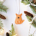 An orange cat-shaped Christmas ornament with a white hanging ribbon, styled on a white background with pine branches, pinecones, and fairy lights.