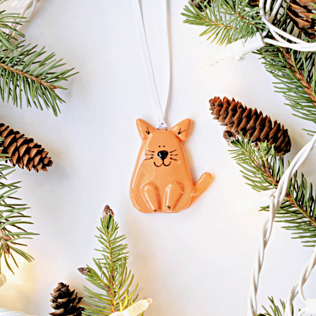 An orange cat-shaped Christmas ornament with a white hanging ribbon, styled on a white background with pine branches, pinecones, and fairy lights.