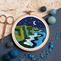 Embroidery hoop with a night scene of cliffs and moonlight, surrounded by embroidery tools on a textured surface.