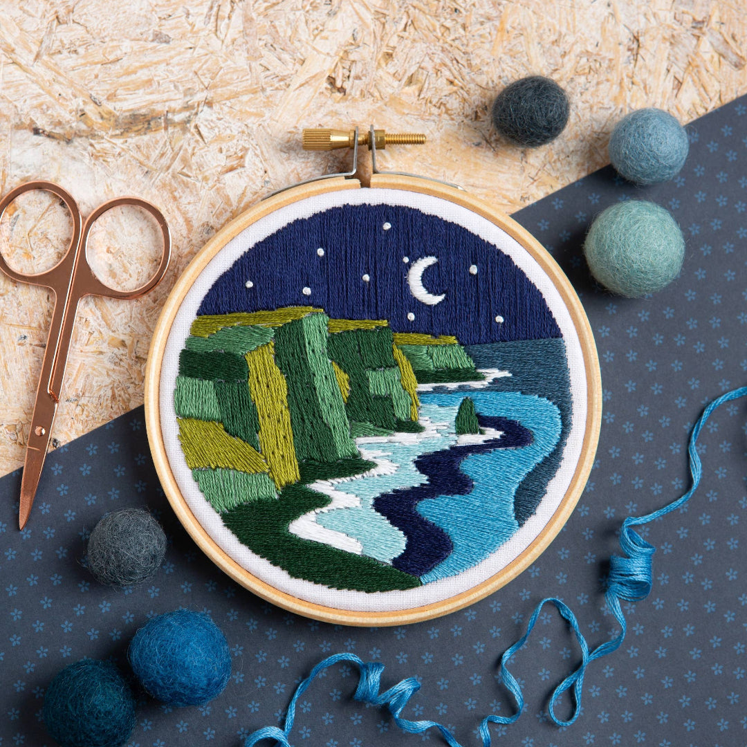 Embroidery hoop with a night scene of cliffs and moonlight, surrounded by embroidery tools on a textured surface.