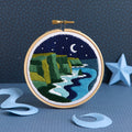 Embroidery hoop with a night scene of cliffs and moon, on a blue textured background.