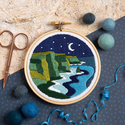 Embroidery hoop with a night scene of cliffs and moonlight, surrounded by embroidery tools on a textured surface.