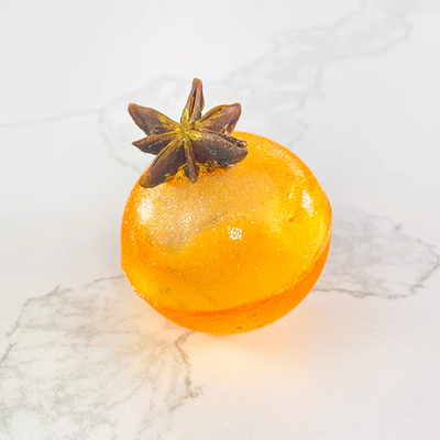 An orange, spherical tea bomb topped with a star anise. The tea bomb is resting on a white marble surface.