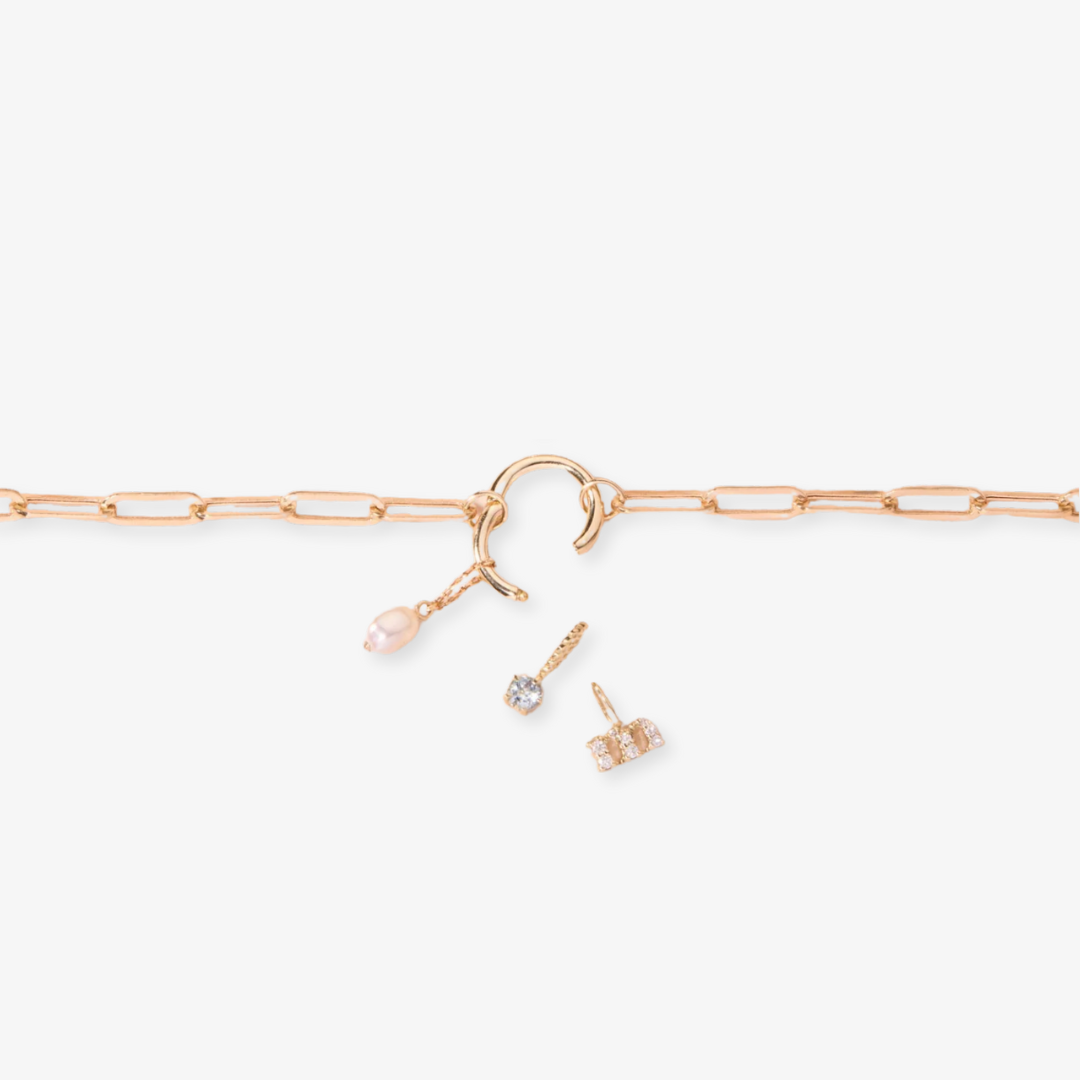 A gold-toned charm necklace with a paper clip link chain, featuring a clasp with attached charms including a white pearl drop and two small crystal pendants, possibly Toniq brand, displayed on a white background. This delicate necklace is available in Ottawa, Ontario, Canada.