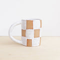 White ceramic mug with brown checkered pattern on a beige surface