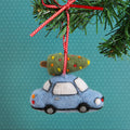 Felt ornament of a blue car carrying a Christmas tree on a green branch with a red ribbon against a teal background.