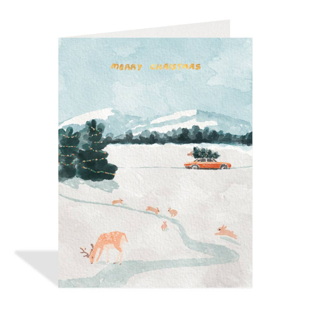 A card with a watercolour illustration of a red car with a tree on the roof driving down a snowy mountain road with deer and bunnies on the side of the road