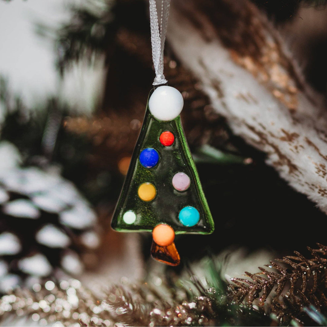 A triangular glass Christmas tree ornament with colorful painted dots and a silver hanging ribbon, photographed hanging in a real decorated tree setting with blurred branches and snow.