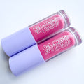 Two tubes of GLOSS by MIMI Coconut Dream Lip Gloss with lavender caps and soft pink shimmer gloss inside, styled to highlight the tropical-inspired cosmetic.