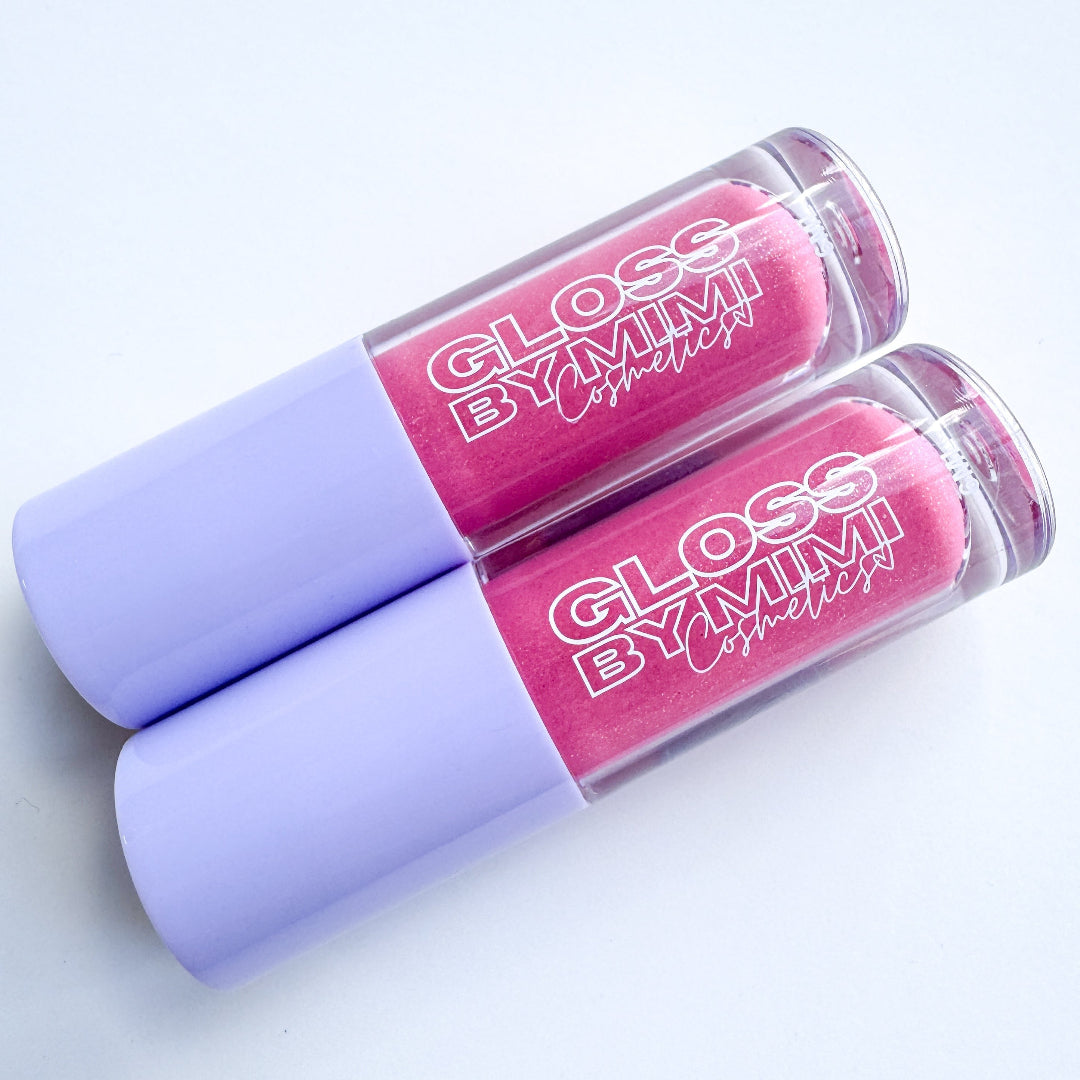 Two tubes of GLOSS by MIMI Coconut Dream Lip Gloss with lavender caps and soft pink shimmer gloss inside, styled to highlight the tropical-inspired cosmetic.