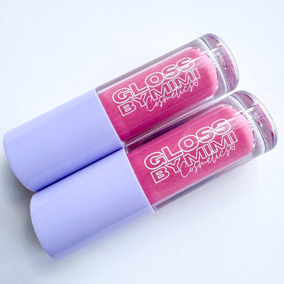 Two tubes of GLOSS by MIMI Coconut Dream Lip Gloss with lavender caps and soft pink shimmer gloss inside, styled to highlight the tropical-inspired cosmetic.