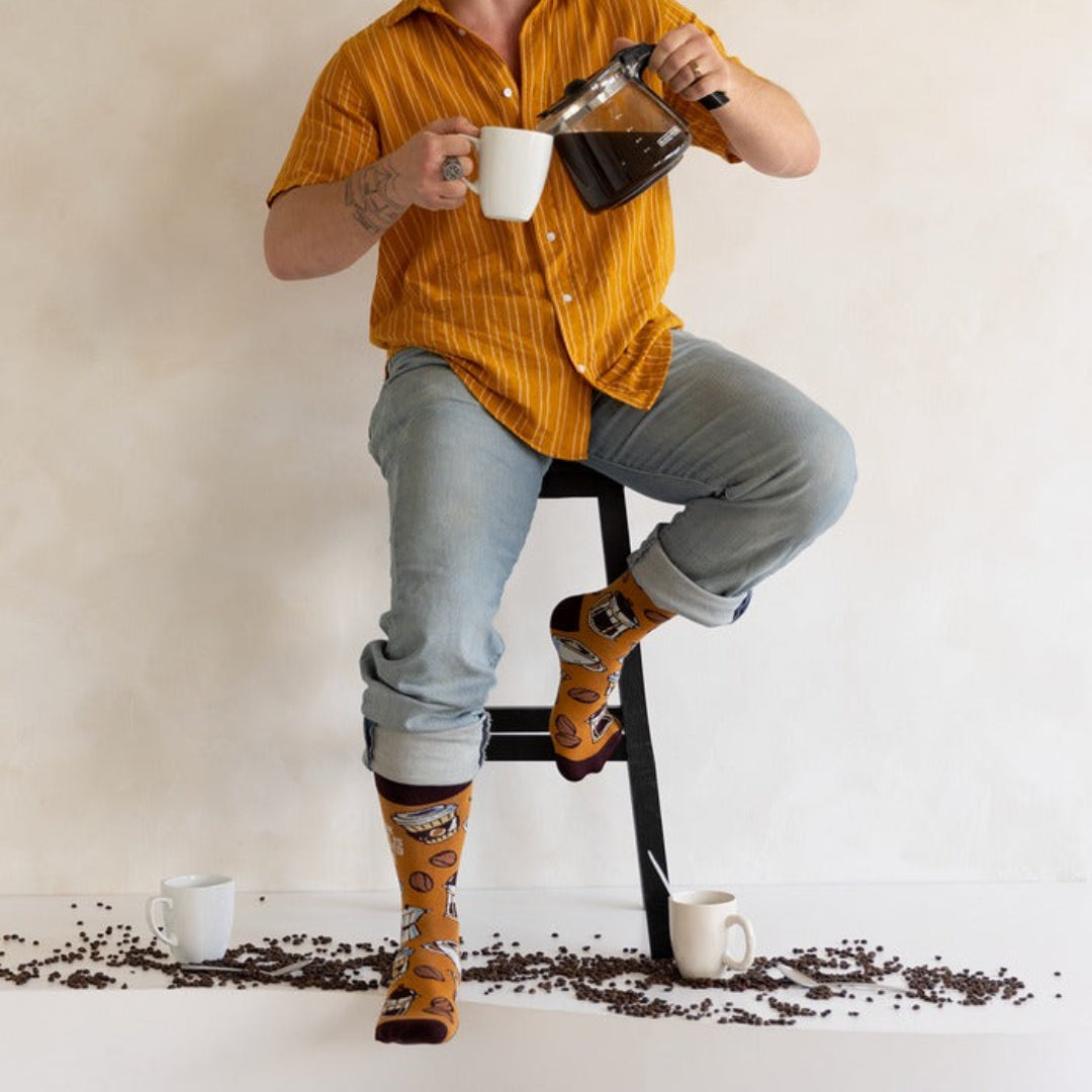 A man sitting on a black stool pours coffee from a glass carafe into mugs while wearing bright yellow striped short-sleeve button-up shirts, light blue jeans, and orange socks with a cartoon character design. Coffee beans are scattered on the floor around him against a textured beige wall.