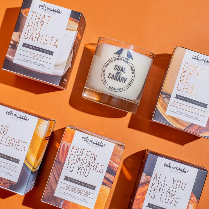 A flat lay of several Coal & Canary candles on an orange background. The candles are in glass jars with gold labels and come in different scents like 
