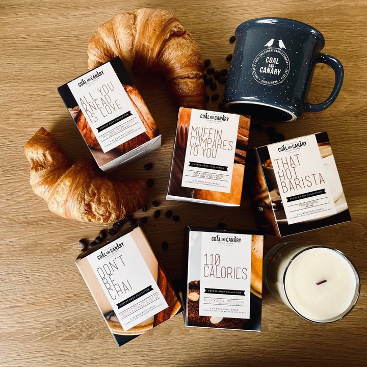 A flat lay of Coal & Canary candles on a wooden table. There are also a mug with the Coal & Canary logo, coffee beans, and a croissant. The candles are packaged in boxes with different scents like 