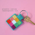An overhead shot of a colourful needlepoint keychain kit on a pink background. A finished square keychain with a patchwork of vibrant colours (pinks, oranges, yellows, greens, blues) is attached to a silver key ring with a brass key.