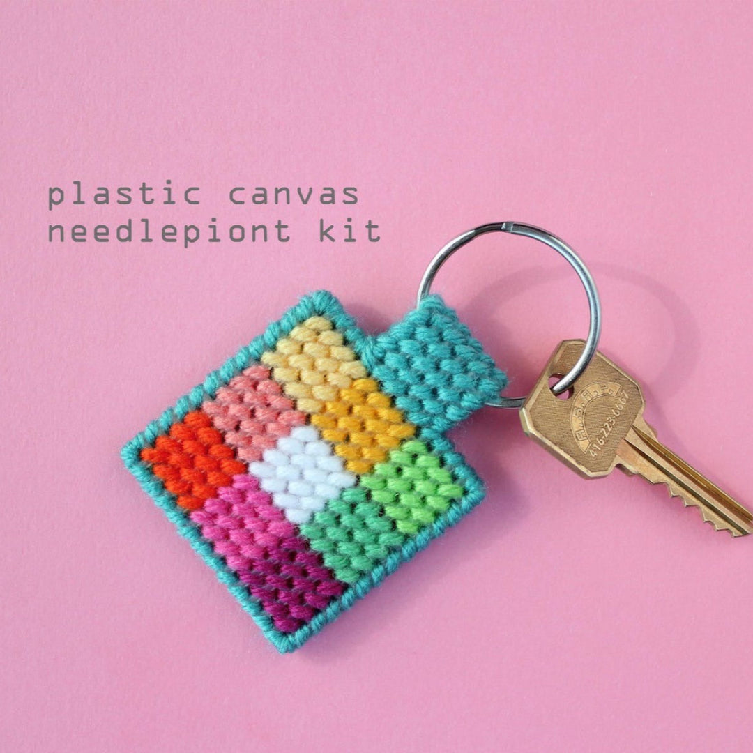 An overhead shot of a colourful needlepoint keychain kit on a pink background. A finished square keychain with a patchwork of vibrant colours (pinks, oranges, yellows, greens, blues) is attached to a silver key ring with a brass key.