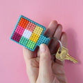 An overhead shot of a colourful needlepoint keychain kit on a pink background. A finished square keychain with a patchwork of vibrant colours (pinks, oranges, yellows, greens, blues) is attached to a silver key ring with a brass key.