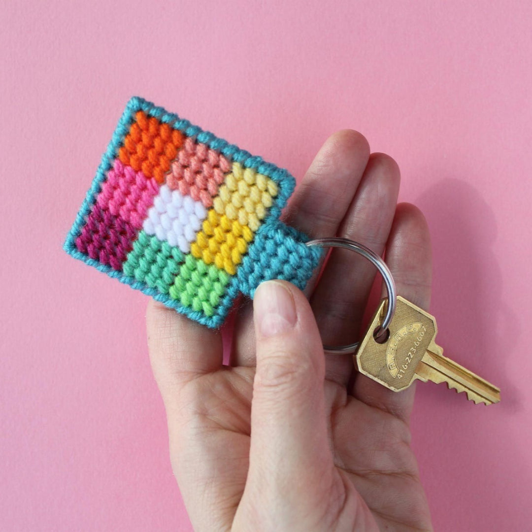 An overhead shot of a colourful needlepoint keychain kit on a pink background. A finished square keychain with a patchwork of vibrant colours (pinks, oranges, yellows, greens, blues) is attached to a silver key ring with a brass key.
