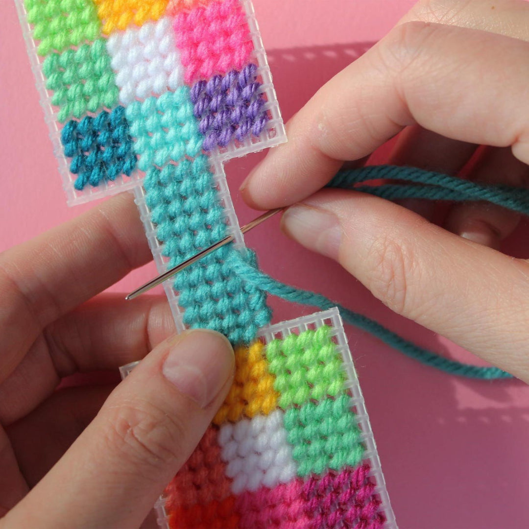 An overhead shot of a colourful needlepoint keychain kit on a pink background. A finished square keychain with a patchwork of vibrant colours (pinks, oranges, yellows, greens, blues) is attached to a silver key ring with a brass key.