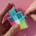 An overhead shot of a colourful needlepoint keychain kit on a pink background. A finished square keychain with a patchwork of vibrant colours (pinks, oranges, yellows, greens, blues) is attached to a silver key ring with a brass key.