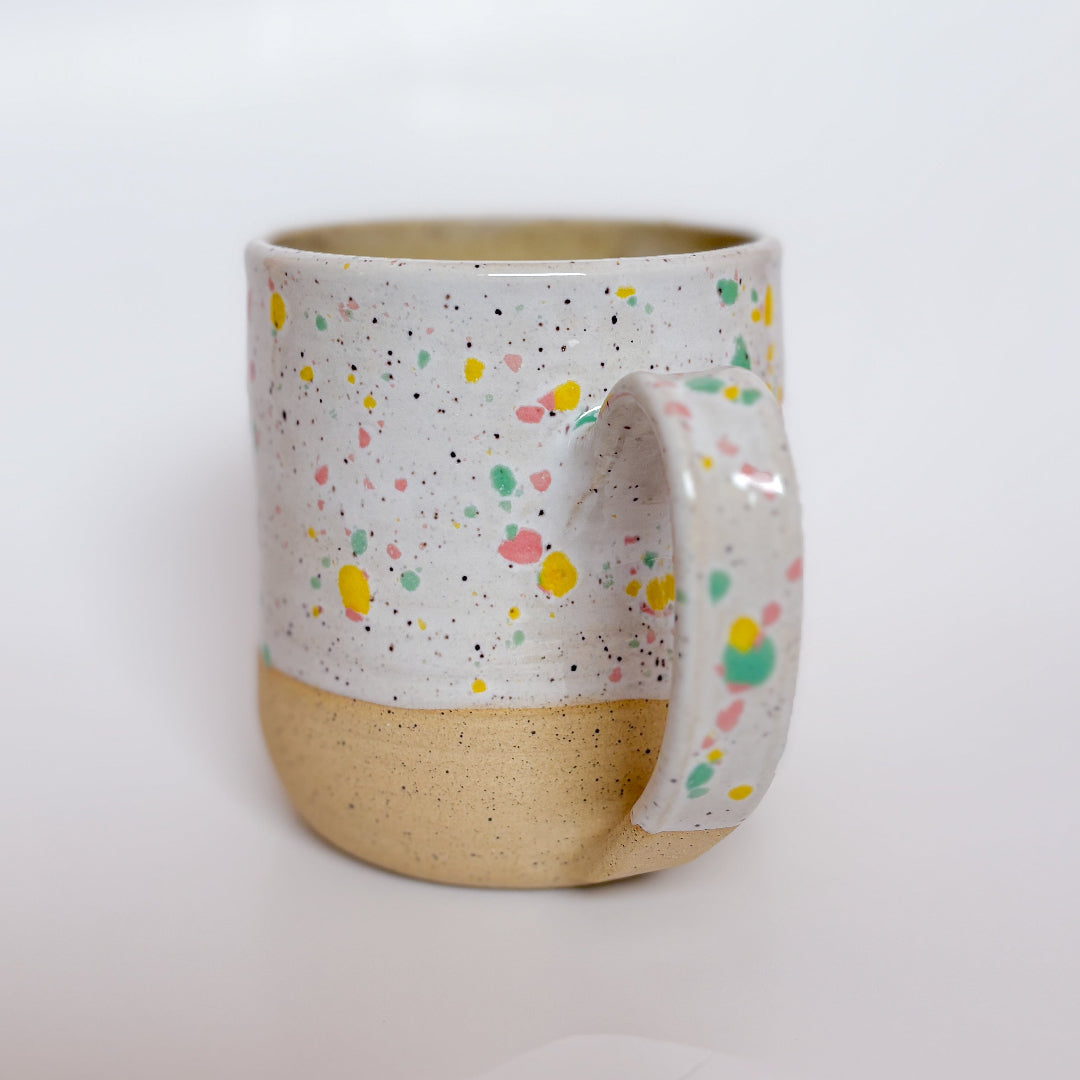 A handmade ceramic mug by Valerie Daoust Ceramics, featuring a white glaze with colorful speckles (pink, yellow, green), displayed on a light gray background. The mug has a large, rounded handle, a speckled texture, and is made in Mississippi Mills, Ontario, Canada.