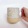 A handmade ceramic mug by Valerie Daoust Ceramics, featuring a white glaze with colorful speckles (pink, yellow, green), displayed on a light gray background. The mug has a large, rounded handle, a speckled texture, and is made in Mississippi Mills, Ontario, Canada.