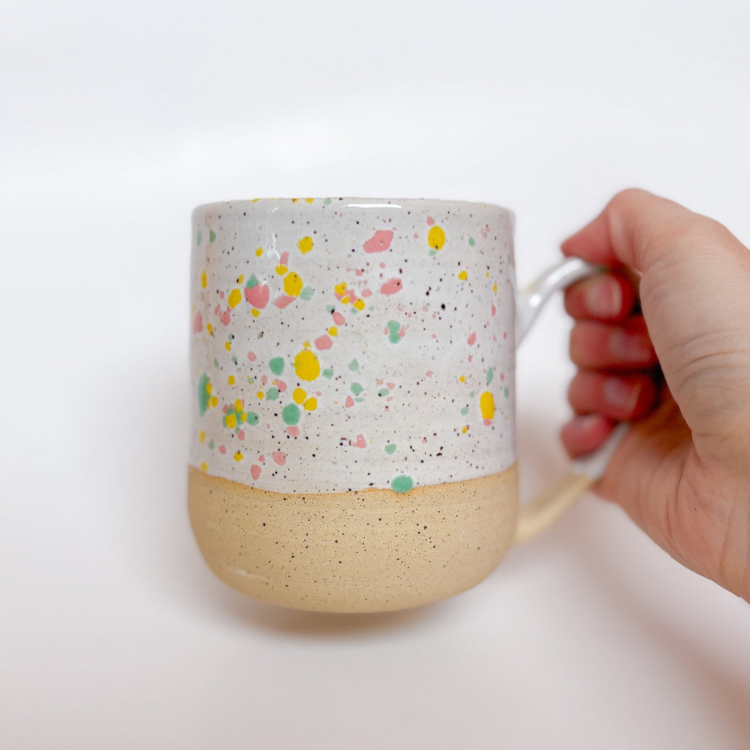 A handmade ceramic mug by Valerie Daoust Ceramics, featuring a white glaze with colorful speckles (pink, yellow, green), displayed on a light gray background. The mug has a large, rounded handle, a speckled texture, and is made in Mississippi Mills, Ontario, Canada.