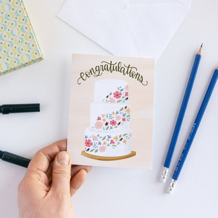 A congratulations card featuring a three-tiered wedding cake decorated with colorful flowers. The word 
