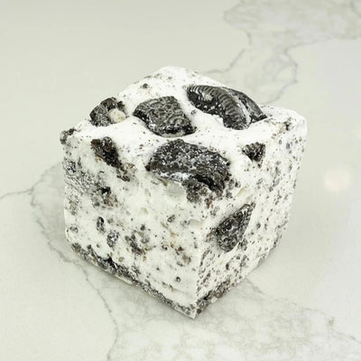 A square marshmallow with swirls of white and black Oreo cookie pieces. The marshmallow is resting on a white marble surface.