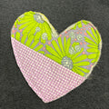 A heart-shaped fabric patch with a pink and white gingham top half and a lime green floral patterned bottom half, placed on a dark gray textured fabric background.