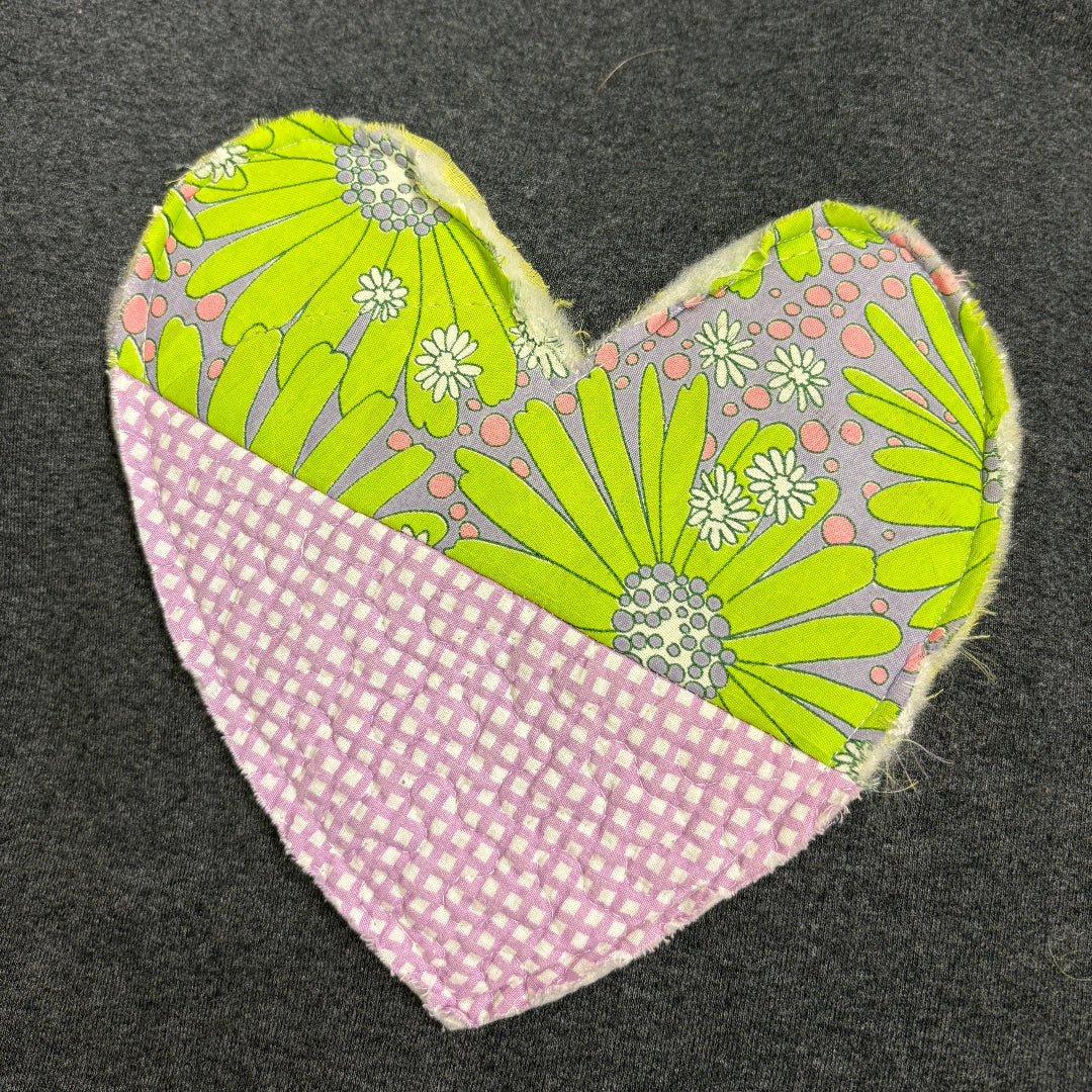 A heart-shaped fabric patch with a pink and white gingham top half and a lime green floral patterned bottom half, placed on a dark gray textured fabric background.
