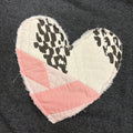 A handmade heart-shaped quilted patch featuring a pink and cream geometric top section and two cream lower sections with black cow-print spots, laid flat on a dark gray fabric background.