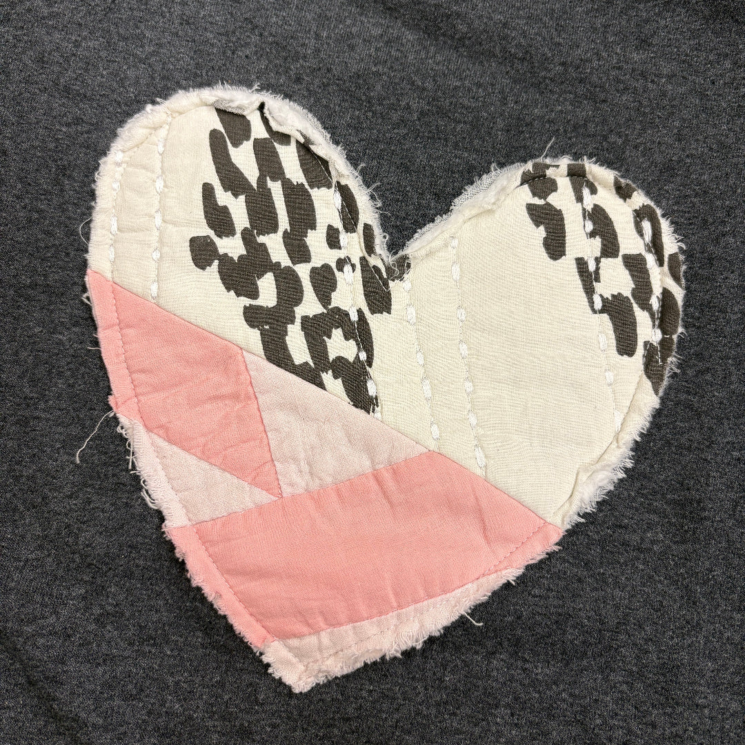 A handmade heart-shaped quilted patch featuring a pink and cream geometric top section and two cream lower sections with black cow-print spots, laid flat on a dark gray fabric background.