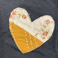 A heart-shaped fabric patch sewn onto a dark gray knit garment. The patch is split diagonally, with one half featuring a cream floral print with pink roses and green leaves, and the other half in a mustard yellow fabric with white dashed stitching.