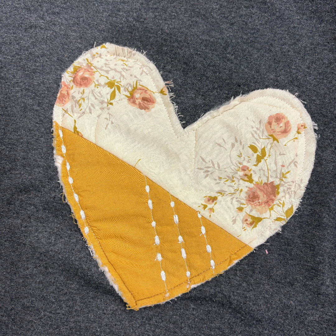 A heart-shaped fabric patch sewn onto a dark gray knit garment. The patch is split diagonally, with one half featuring a cream floral print with pink roses and green leaves, and the other half in a mustard yellow fabric with white dashed stitching.