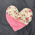 A heart-shaped quilted oven mitt with a cream floral print and a solid pink corner, laid flat on a dark gray fabric background.