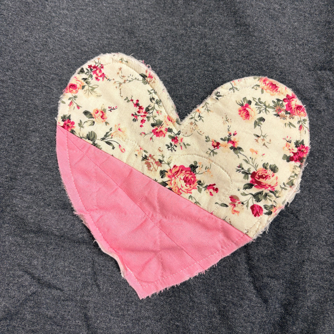 A heart-shaped quilted oven mitt with a cream floral print and a solid pink corner, laid flat on a dark gray fabric background.
