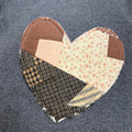 A heart-shaped patchwork quilted fabric applique with various patterned sections, including plaid, floral, and solid brown, displayed on a dark gray textured garment.
