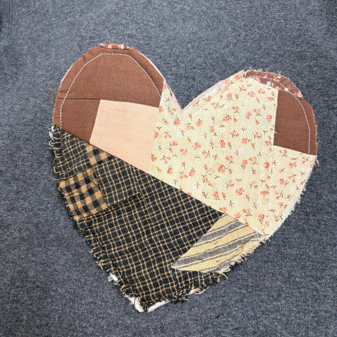 A heart-shaped patchwork quilted fabric applique with various patterned sections, including plaid, floral, and solid brown, displayed on a dark gray textured garment.