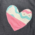 A close-up of a dark gray heathered sweatshirt featuring a large, handmade-style quilted heart patch on the chest. The heart is split diagonally, with one half in solid pink and the other half in a wavy pattern of white, light blue, and pink.