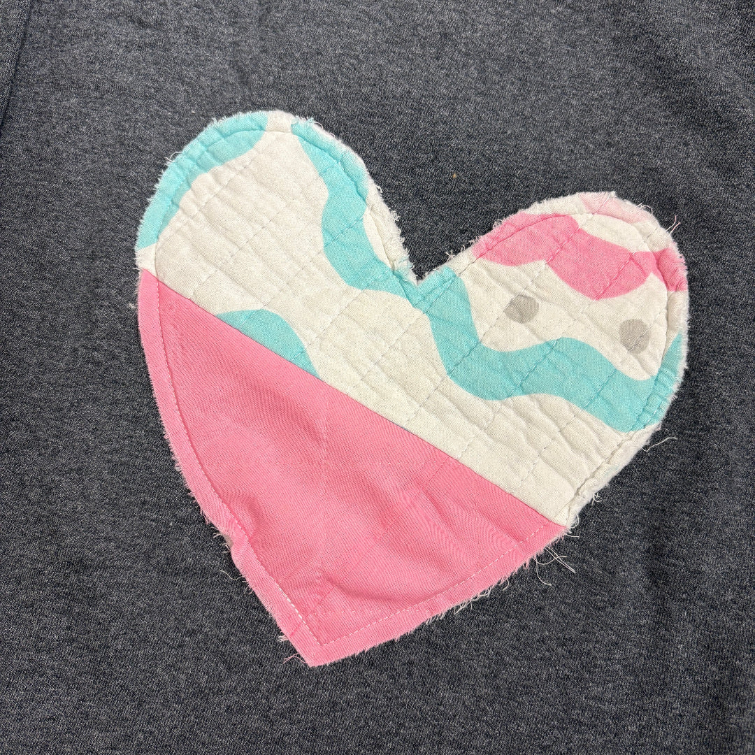 A close-up of a dark gray heathered sweatshirt featuring a large, handmade-style quilted heart patch on the chest. The heart is split diagonally, with one half in solid pink and the other half in a wavy pattern of white, light blue, and pink.