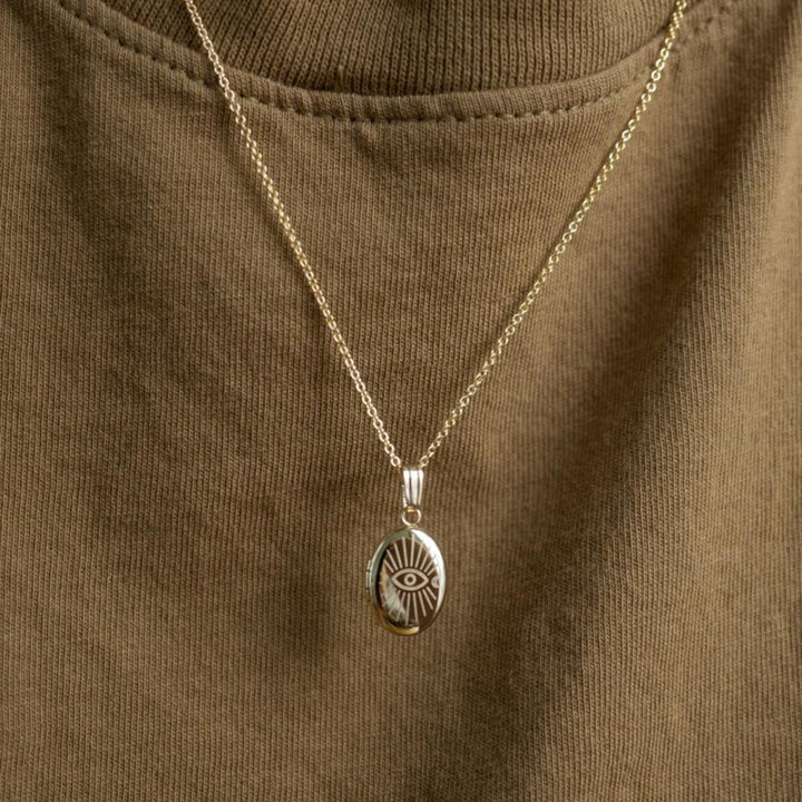 A copper oval locket necklace with an engraved evil eye design, featuring an eye with radiating lines and crescent moons, hanging from a gold chain. This is a protective and spiritual piece of jewelry, available in Ottawa, Ontario, Canada.