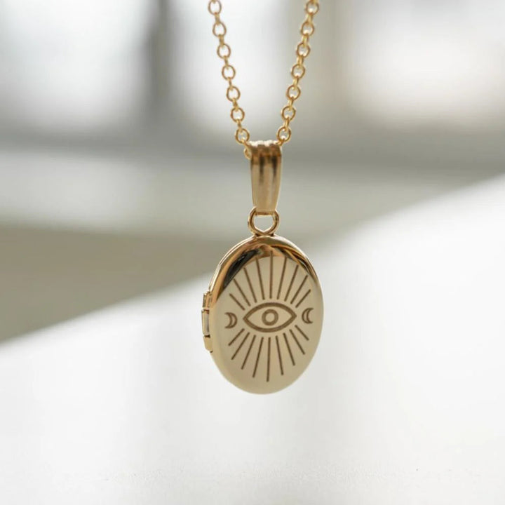 A copper oval locket necklace with an engraved evil eye design, featuring an eye with radiating lines and crescent moons, hanging from a gold chain. This is a protective and spiritual piece of jewelry, available in Ottawa, Ontario, Canada.