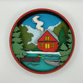 Decorative wall piece with a red cabin, trees, and water on a white background