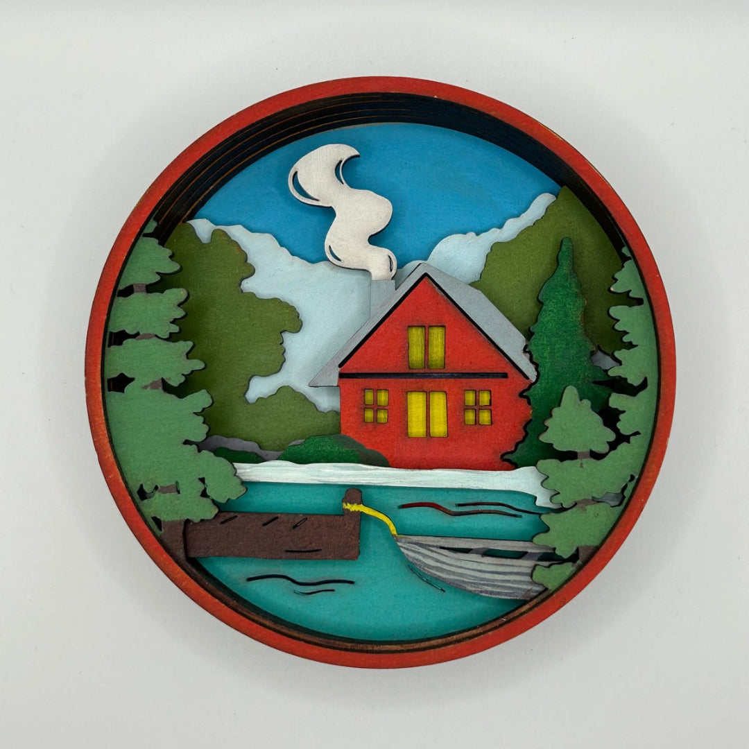 Decorative wall piece with a red cabin, trees, and water on a white background