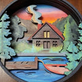 Wooden cutout of a cabin by a lake with trees and mountains at sunset.