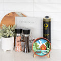 A styled kitchen countertop scene featuring a round illustrated coaster on a small stand, two black pepper and salt shakers, a tall black bottle of olive oil, a white book, and a potted plant against a white subway tile background.