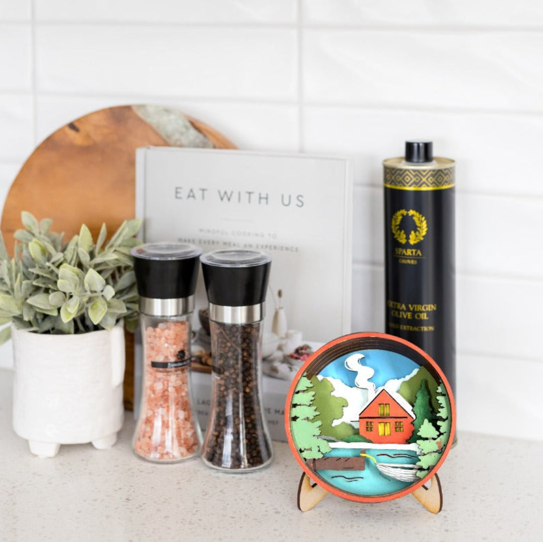 A styled kitchen countertop scene featuring a round illustrated coaster on a small stand, two black pepper and salt shakers, a tall black bottle of olive oil, a white book, and a potted plant against a white subway tile background.