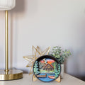 A round decorative ceramic clock with a colorful lakeside cabin scene, displayed on a tabletop next to a gold lamp base, a star-shaped decor object, and a small potted plant.